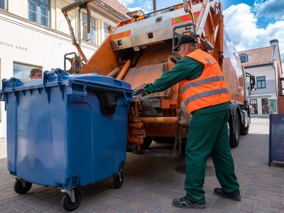 Oranžā atstarojošā vestē tērpies SIA Kuldīgas komunālie sētnieks stumj lielu zilu atkritumu konteineru pie oranžas atkritumu savākšanas mašīnas. Fonā redzamas vecpilsētas ēkas.