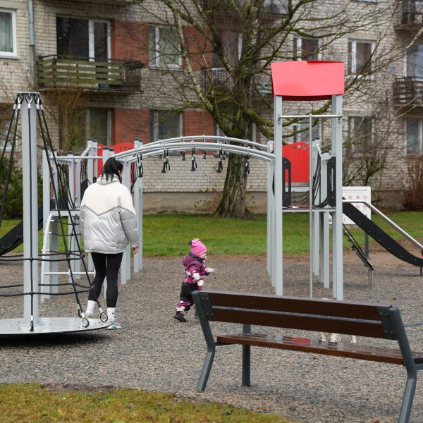 Sieviete sudraba virsjakā un mazs bērns rozā cepurē un violetā kombinezonā atrodas modernā rotaļu laukumā. Sieviete stāv pie rotējoša karuseļa, kamēr bērns iet viņas virzienā. Fonā redzama daudzdzīvokļu māja.