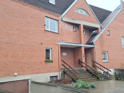 Sarkana ķieģeļu ēka ar stāviem jumtiem un dažādu formu logiem. Galvenā ieeja ar koka margām ved uz durvīm zem nelielas nojumes.