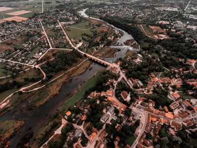 Gaisa skats uz Kuldīgas pilsētu ar Ventas upi, veco ķieģeļu tiltu un plašo Ventas rumbu. Abpus upei redzamas ēkas ar sarkanīgiem jumtiem, ko ieskauj zaļi lauki un koki.