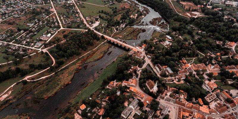 Gaisa skats uz Kuldīgas pilsētu ar Ventas upi, veco ķieģeļu tiltu un plašo Ventas rumbu. Abpus upei redzamas ēkas ar sarkanīgiem jumtiem, ko ieskauj zaļi lauki un koki.