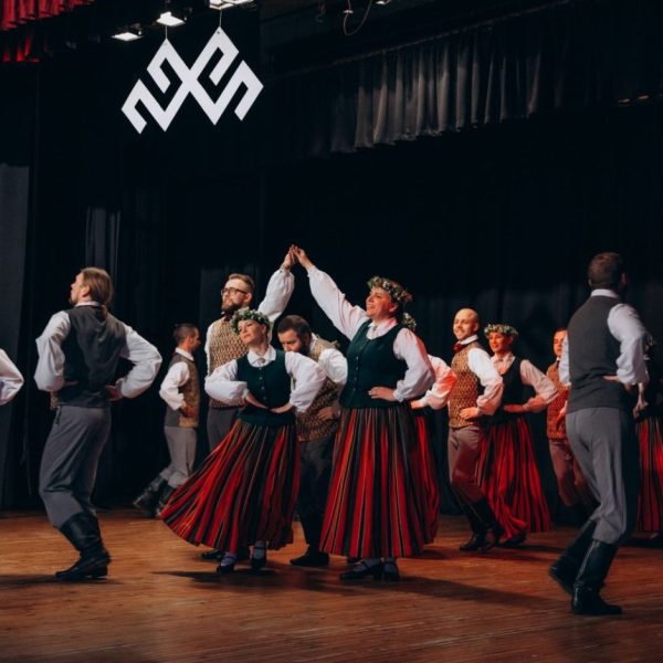 Folkloras dejotāji tradicionālos latviešu tērpos uzstājas uz skatuves. Sievietes valkā garus svītrainus svārkus, zaļas vestes un vainagus, vīrieši – pelēkas bikses un vestes. Aiz dejotājiem pie melnās sienas redzama liela balta Jumja zīme.