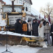 Grupa cilvēku, tostarp jaunieši, ziemas apģērbā stāv sniegotā laukumā pie liela dzeltena sniega tīrītāja ar uzgriezni priekšā. Daži no viņiem tur Latvijas karogus. Uz sniega tīrītāja ir zīme ar tekstu "LABĀK MANU GALVU ŅĒMA N MANU".