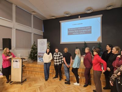 Sieviete rozā džemperī runā no pults auditorijas priekšā, kurā stāv vairākas sievietes un vīrieši.