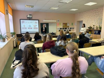 Klasē pie prožektora ekrāna, kurā redzams uzraksts "Ar ko sākt?", "Ideja", "Tēma", stāv sieviete, prezentējot studentiem, kas sēž pie galdiem. Otra sieviete stāv klases labajā pusē.