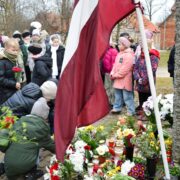 Bērni noliek ziedus un iedegtas sveces pie akmens pieminekļa, kura pakājē jau izvietoti daudzi ziedi un svecītes. Blakus plīvo Latvijas karogs. Fonā stāv vēl citi bērni un pieaugušie. Komunistiskā genocīda upuru piemiņas pasākumi novadā
