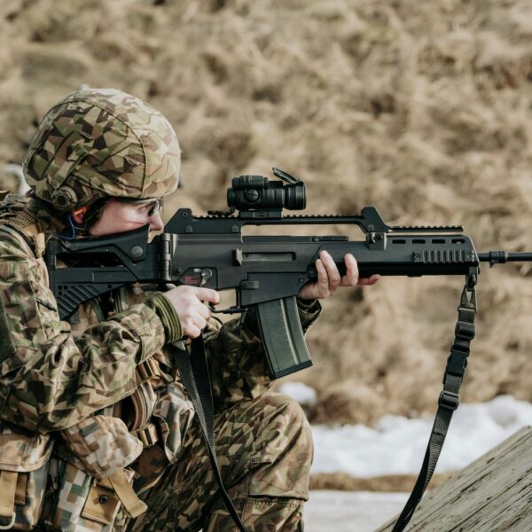 Notupies karavīrs kamuflāžas uniformā ar Latvijas karoga plāksteri un ķiveri mērķē ar šauteni ar optisko tēmekli, valkājot dzirdes aizsargus.